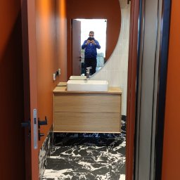 Usługi Ogólnobudowlane Kamil Szustek - Nowoczesna łazienka z umywalką na drewnianej szafce, marmurową podłogą i pomarańczowymi ścianami. W lustrze odbija się osoba robiąca zdjęcie.