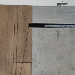 Usługi Ogólnobudowlane Kamil Szustek - Układanie paneli podłogowych w trakcie prac. Widoczne panele z wzorem drewna, listwa dylatacyjna i surowy beton. Na górze widoczne płytki na suficie z krzyżykami.