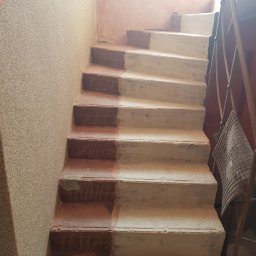 Usługi Ogólnobudowlane Kamil Szustek - Schody w trakcie renowacji: połowa stopni pomalowana na biało, druga w kolorze ceglastym. Ściana z fakturą, widoczne metalowe elementy balustrady.