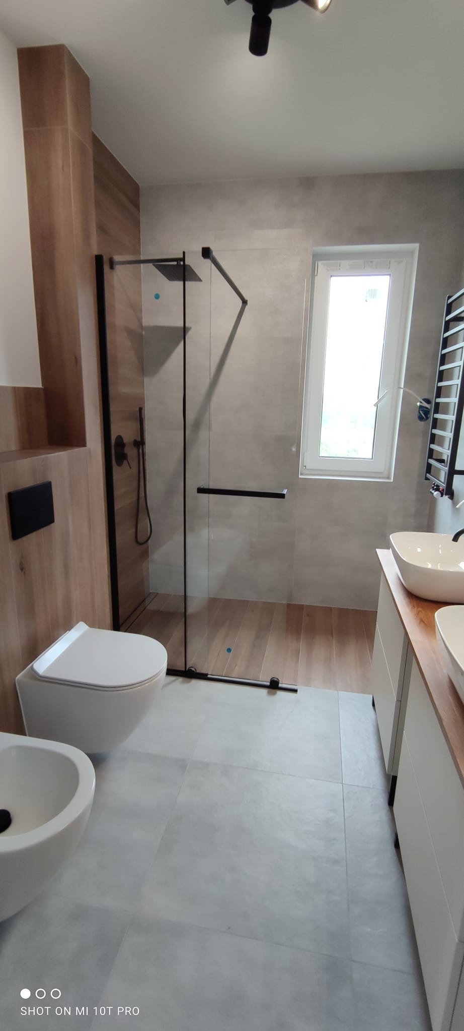 Nowoczesna łazienka z kabiną prysznicową typu walk-in z czarnymi ramami, wiszącą toaletą i bidetem. Umywalki nablatowe na drewnianym blacie. Szare płytki i drewniane akcenty.
