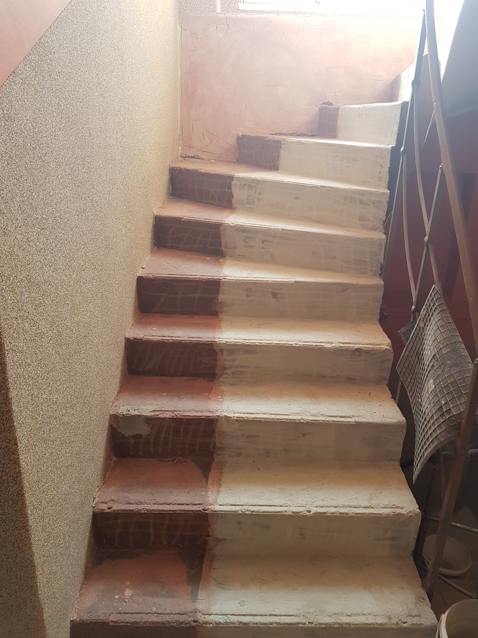 Schody w trakcie renowacji: połowa stopni pomalowana na biało, druga w kolorze ceglastym. Ściana z fakturą, widoczne metalowe elementy balustrady.