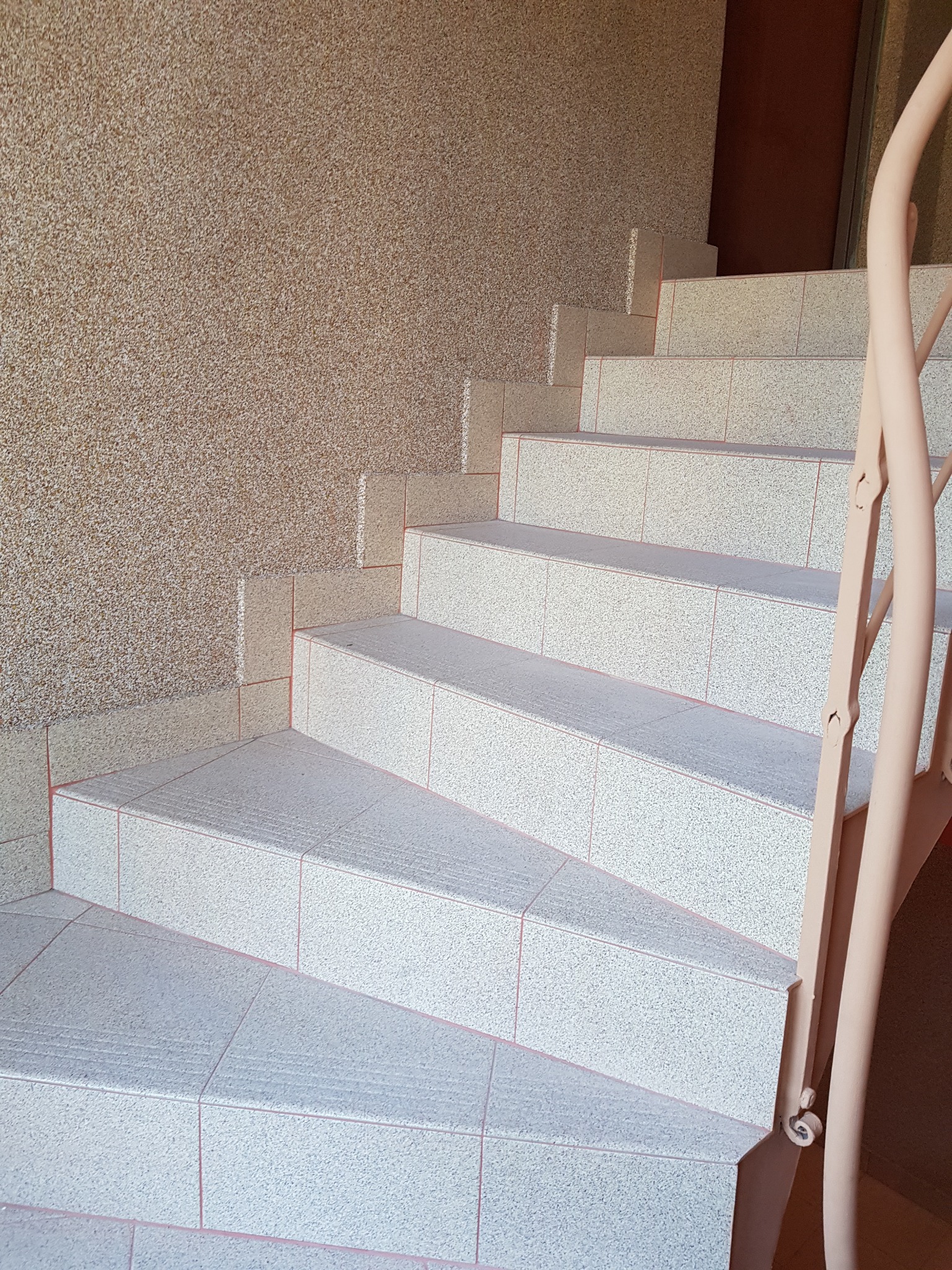 Schody wyłożone jasnymi płytkami z delikatnym wzorem, z beżową balustradą i ścianą o fakturze baranka. Widoczne fugi w kolorze różowym.