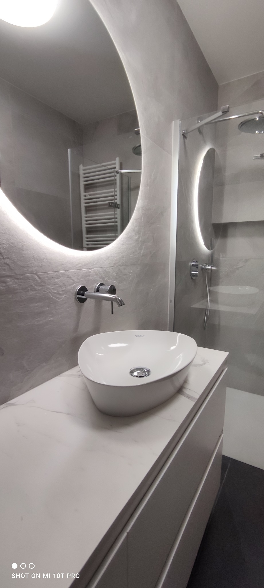 Nowoczesna łazienka w szarościach z owalną umywalką nablatową, podświetlanym lustrem i kabiną prysznicową. Minimalistyczny design, marmurowy blat.