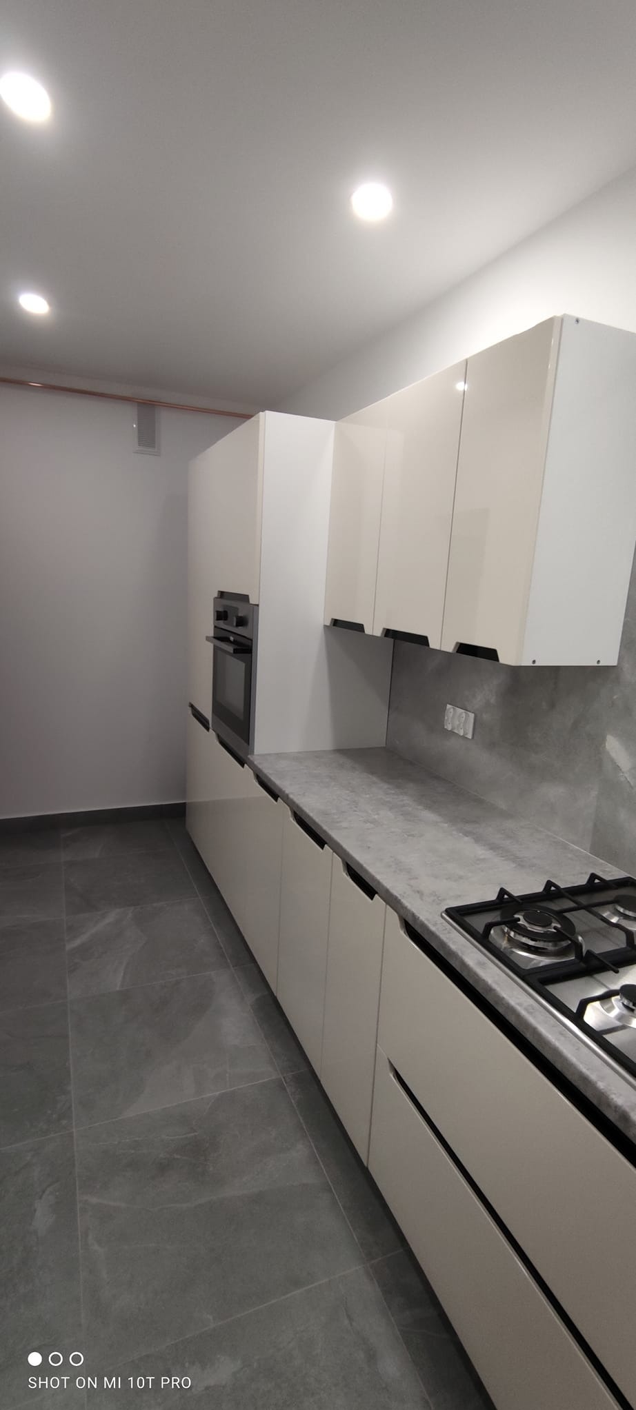 Nowoczesna kuchnia z białymi szafkami, szarym blatem i płytkami podłogowymi. Widoczna płyta gazowa i piekarnik. Minimalistyczny design, oświetlenie punktowe.