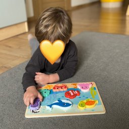 Wioska Szafirowa Agata Stańczyk - Chłopiec leżący na dywanie układa drewniane puzzle z motywem morskim, ćwicząc koordynację wzrokowo-ruchową i rozpoznawanie kształtów.