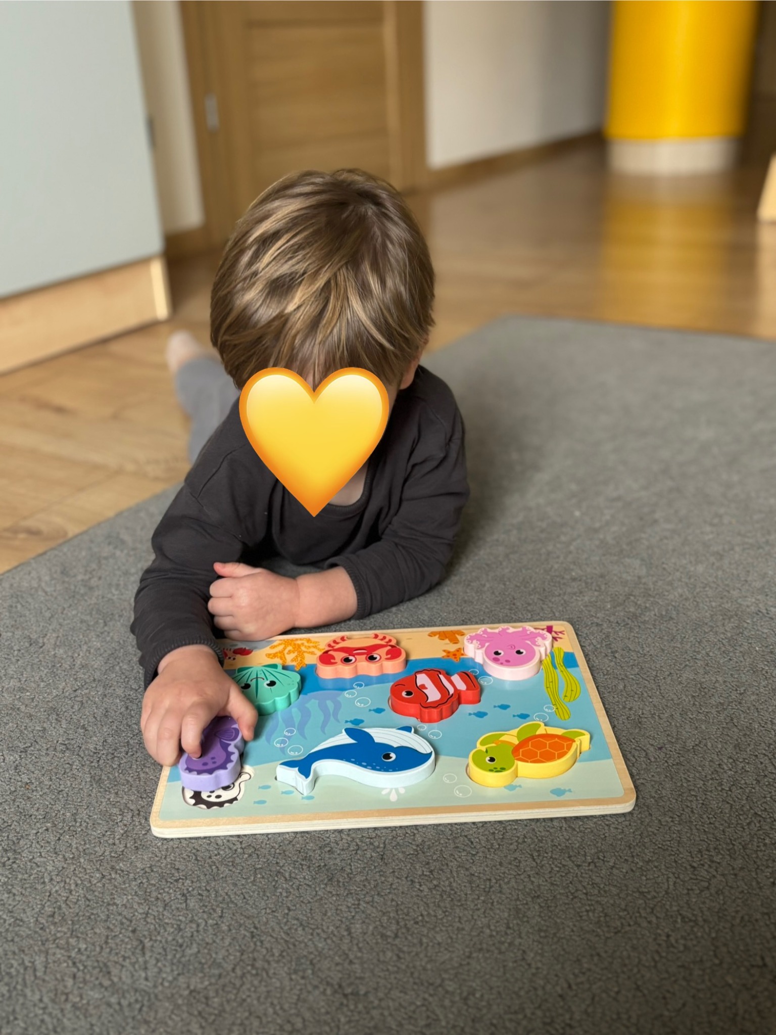 Chłopiec leżący na dywanie układa drewniane puzzle z motywem morskim, ćwicząc koordynację wzrokowo-ruchową i rozpoznawanie kształtów.