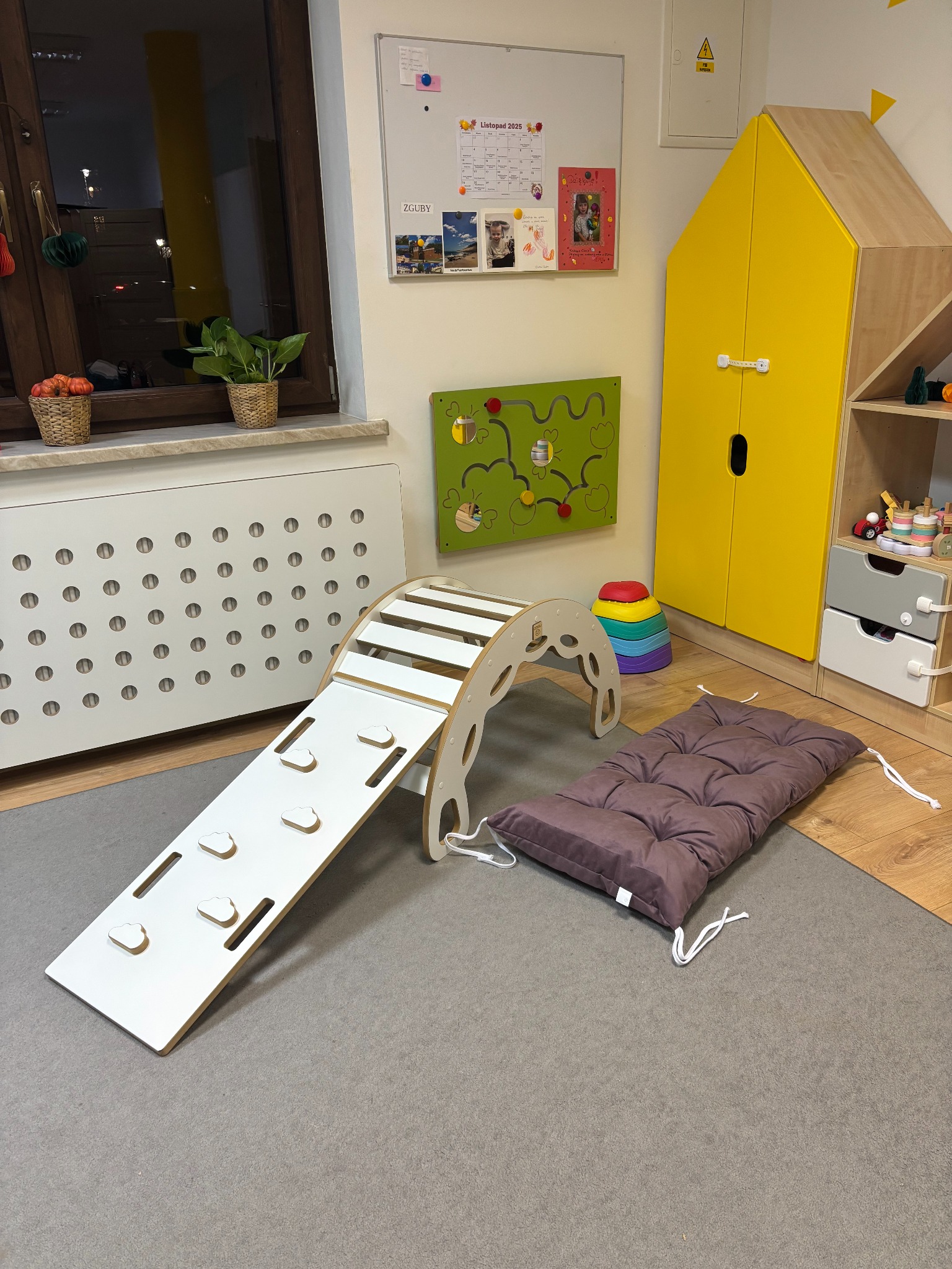 Kącik zabaw w żłobku: drewniany domek-szafa, drabinka wspinaczkowa z poduszką, labirynt na ścianie i kalendarz. Jasne, przytulne wnętrze.
