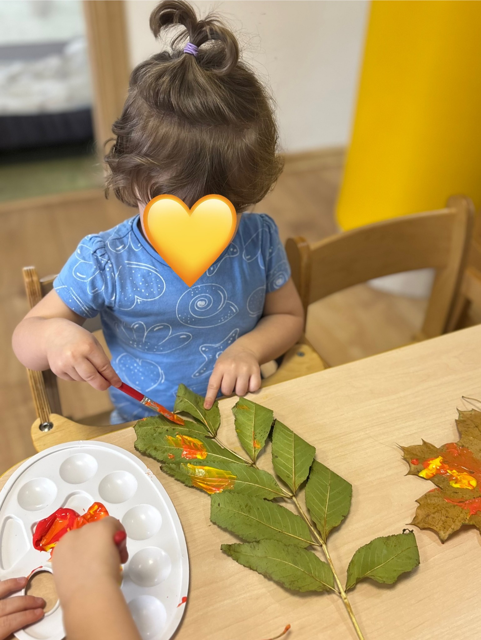 Dziewczynka w błękitnej koszulce maluje pędzlem liście na stole. Widoczna paleta z farbami i fragment dłoni drugiej osoby. Kreatywne zajęcie plastyczne.