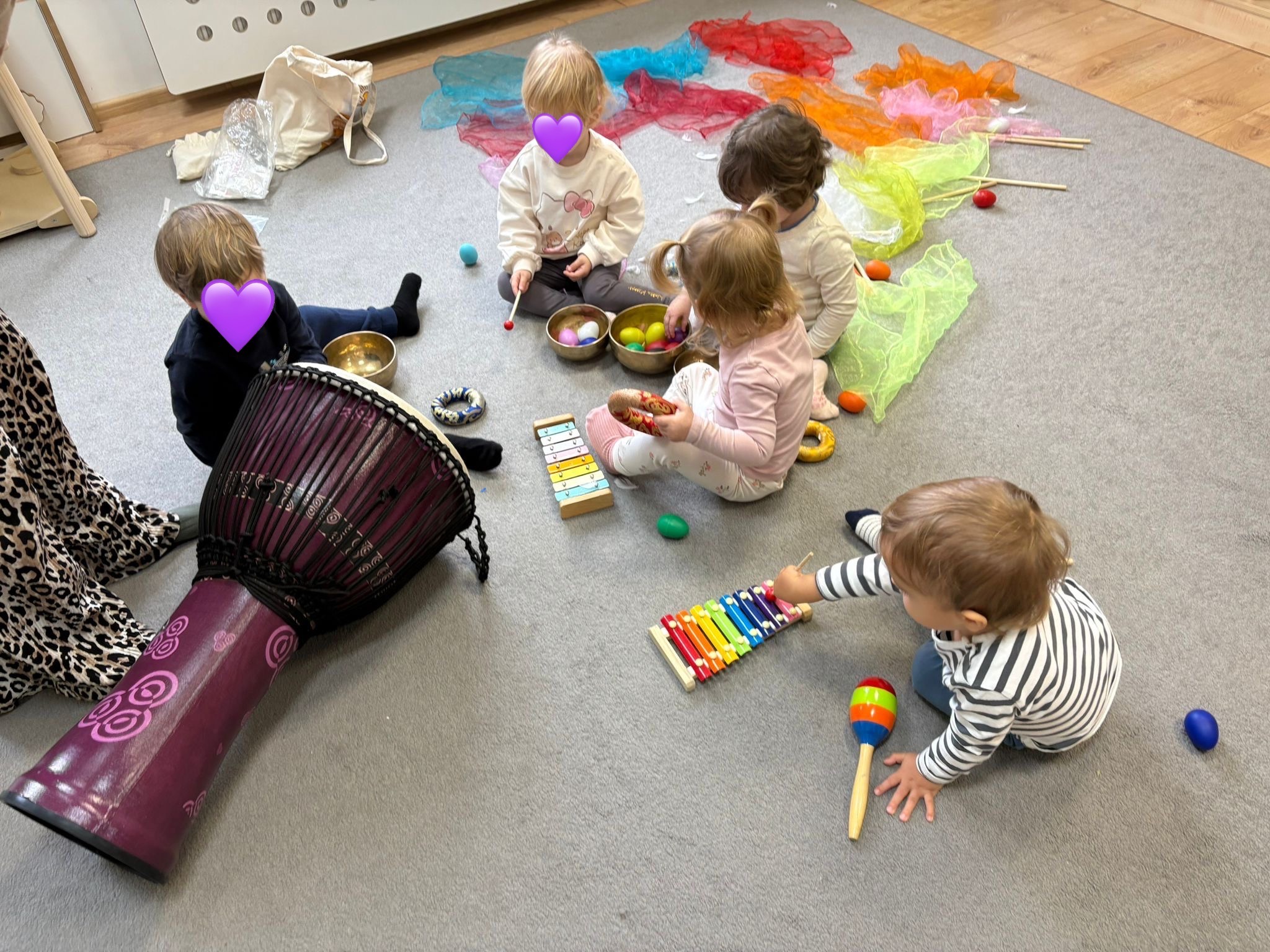 Grupa dzieci siedzi na dywanie, bawiąc się instrumentami muzycznymi: bęben djembe, metalofon, marakasy i misy dźwiękowe. Kolorowe chusty w tle.