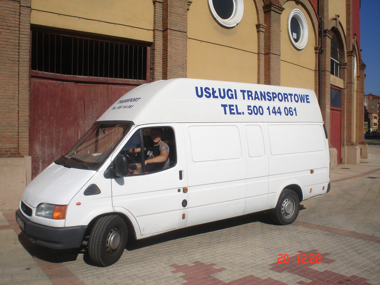 Biały bus z napisem 'Usługi Transportowe' i numerem telefonu zaparkowany na chodniku z czerwonej kostki brukowej przed budynkiem z cegły.