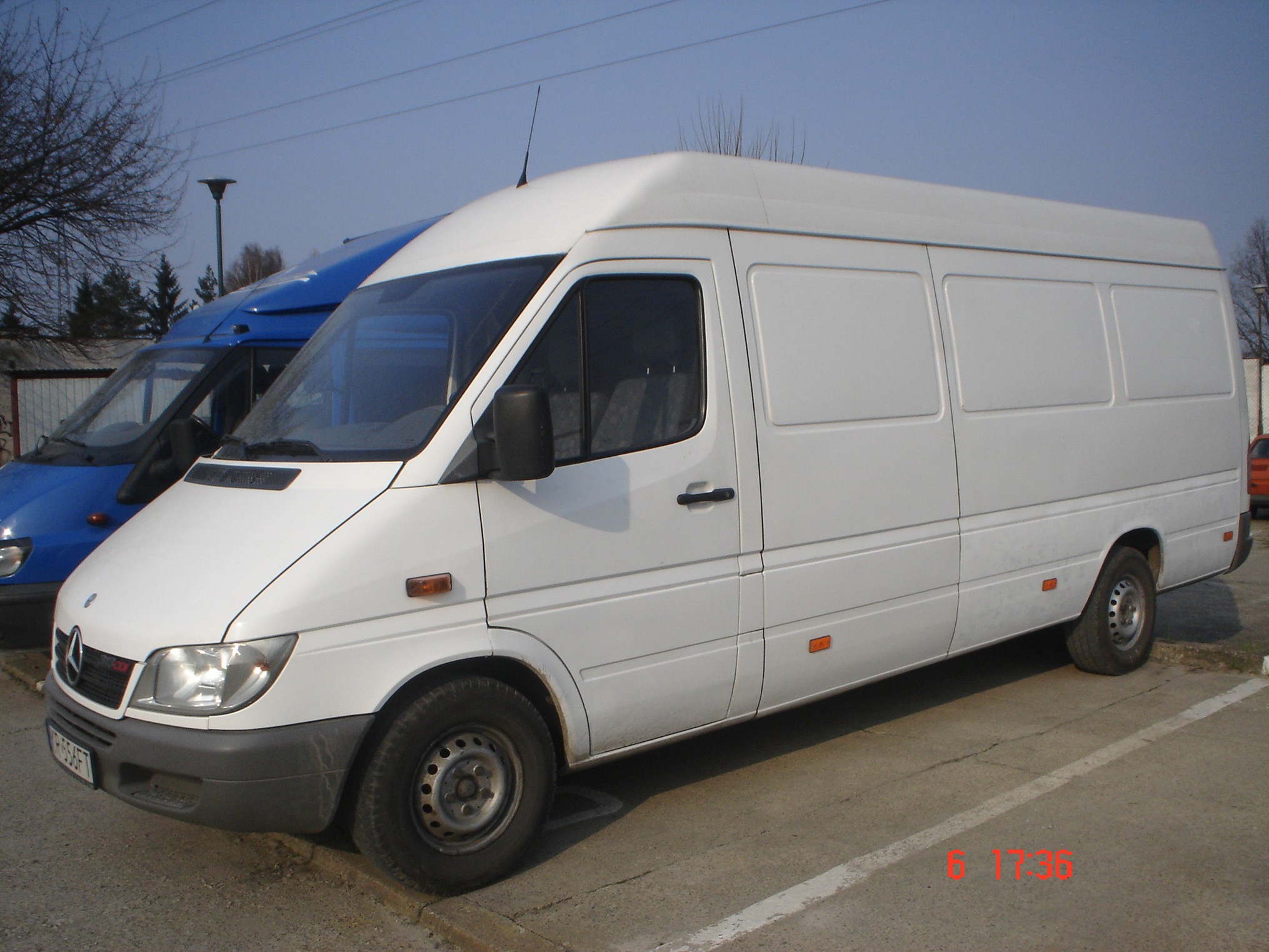 Biały, wysoki furgon dostawczy Mercedes-Benz Sprinter zaparkowany na betonowym placu, widoczny z boku, z niebieskim busem w tle i nagim drzewem po lewej stronie.