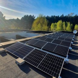 Nobisend - Rząd paneli słonecznych na płaskim dachu budynku, z zielenią drzew w tle i błękitnym niebem. Widoczny satelita na dachu. Słoneczny dzień.