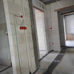 Nobisend SMART System - Układanie instalacji elektrycznej w surowym wnętrzu z jasnych bloczków, z licznymi peszlami na podłodze i czerwonymi puszkami pod osprzęt.