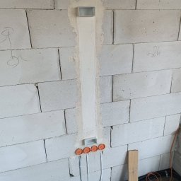 Nobisend SMART System - Montaż okablowania elektrycznego w bruzdach ściany z bloczków, z trzema okrągłymi puszkami i jedną prostokątną, oraz peszlami na podłodze. Widoczne rysunki kredą.