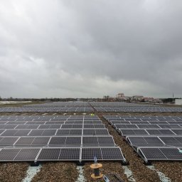 Nobisend SMART System - Rozległa instalacja paneli fotowoltaicznych na płaskim dachu pokrytym żwirem, z rzędami ciemnych modułów i pracownikiem w żółtej kamizelce, pod pochmurnym niebem.