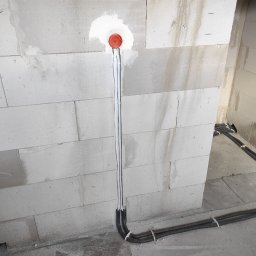 Nobisend SMART System - Instalacja elektryczna w surowym wnętrzu: czerwona puszka w szarej ścianie z bloczków, z której wychodzą białe peszle, łączące się z czarnymi rurami karbowanymi ułożonymi przy podłodze.