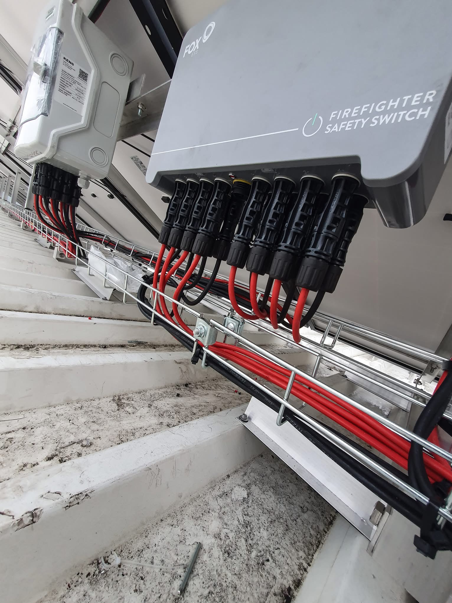 Szary przełącznik bezpieczeństwa przeciwpożarowego Fox ESS z podłączonymi czerwonymi i czarnymi kablami, zamontowany na metalowej konstrukcji na zewnątrz budynku.