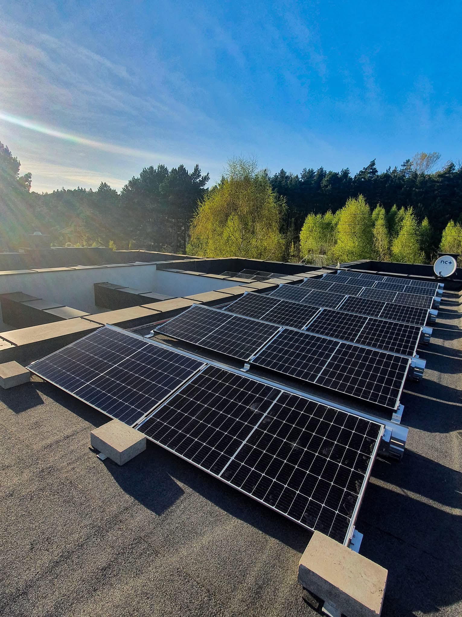 Rząd paneli słonecznych na płaskim dachu budynku, z zielenią drzew w tle i błękitnym niebem. Widoczny satelita na dachu. Słoneczny dzień.