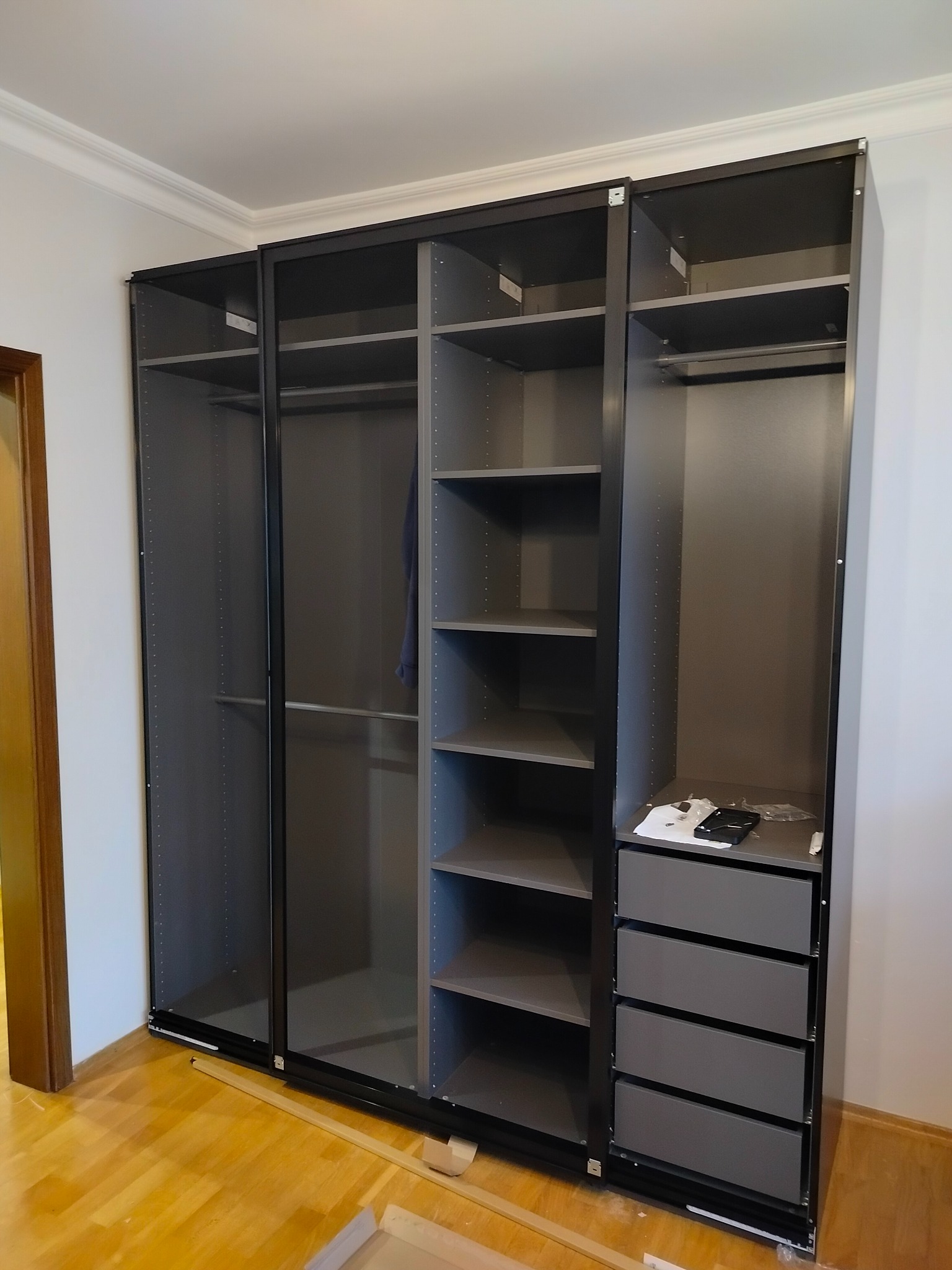 Szara, modułowa szafa z przesuwnymi drzwiami i szufladami, częściowo zmontowana, z widocznymi elementami montażowymi na podłodze. Minimalistyczny design.