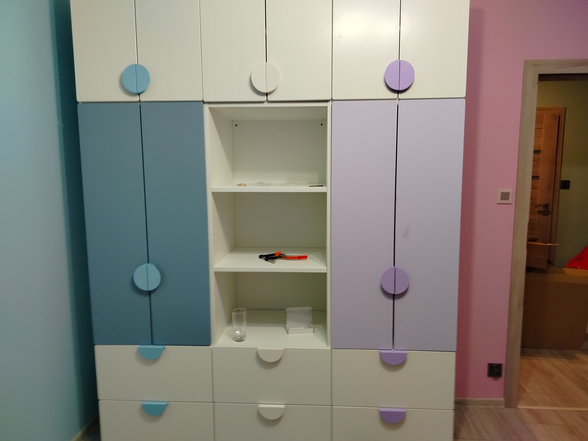 Wielobarwna szafa z okrągłymi uchwytami: błękitne, białe i fioletowe fronty. Na półce leżą narzędzia, a obok stoi szklanka. Widoczny fragment różowej ściany.