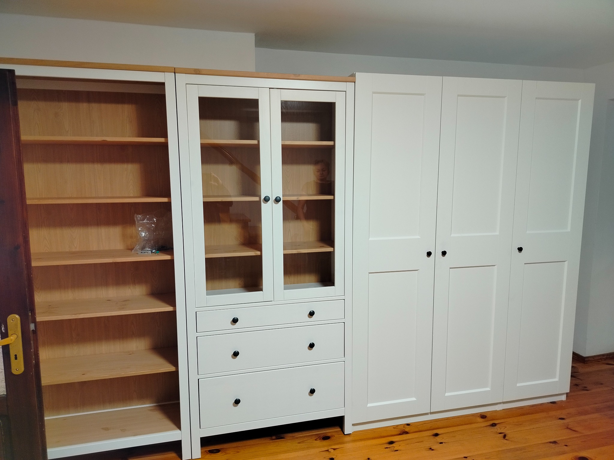 Biały zestaw mebli modułowych z drewnianymi akcentami, obejmujący szafę z przeszklonymi drzwiami, szuflady i wysokie szafki, na tle drewnianej podłogi.