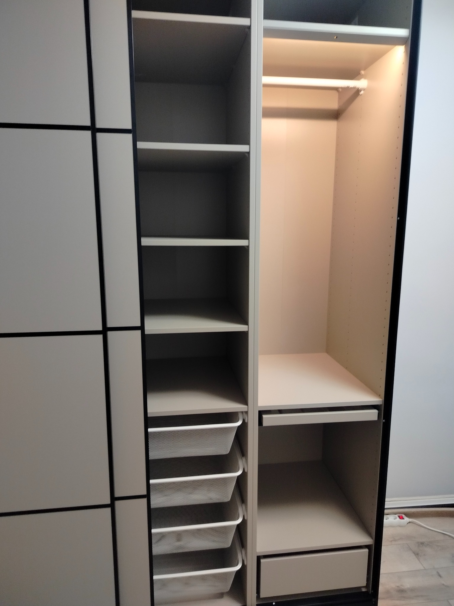 Wnętrze szafy wnękowej z oświetleniem LED, półkami, koszami drucianymi i szufladą. Drzwi przesuwne z podziałem na kwadraty. Widoczny fragment podłogi.