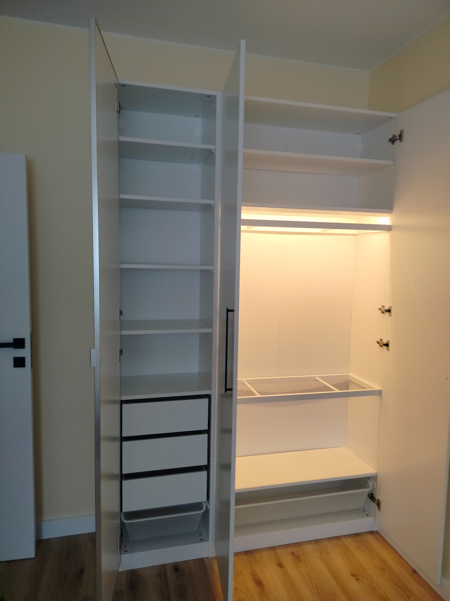 Biała szafa wnękowa z otwartymi drzwiami, widoczne półki i szuflady, oświetlenie LED wewnątrz. Montaż mebli w Justynowie.