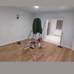 Jacek Jeż - Wnętrze po malowaniu: jasne ściany, drewniana podłoga, drabina z kurtką, widok na sąsiednie pomieszczenie. Świeżo odmalowane mieszkanie.