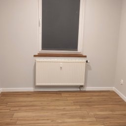 Jacek Jeż - Jasne wnętrze pokoju po malowaniu: białe ściany, drewniana podłoga, okno z roletą i grzejnik. Minimalistyczny wystrój, świeżo po remoncie.