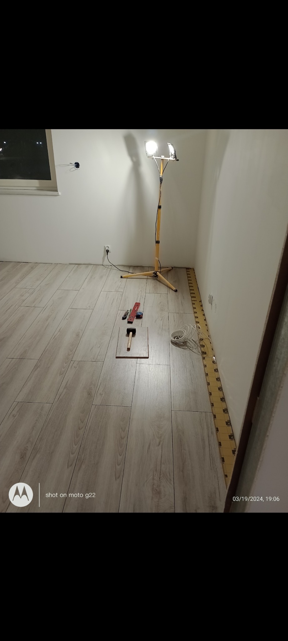 Układanie paneli podłogowych w jasnym wnętrzu. Widoczne narzędzia: młotek, miarka, nóż. Dodatkowe oświetlenie halogenowe. Listwy przypodłogowe w trakcie montażu.