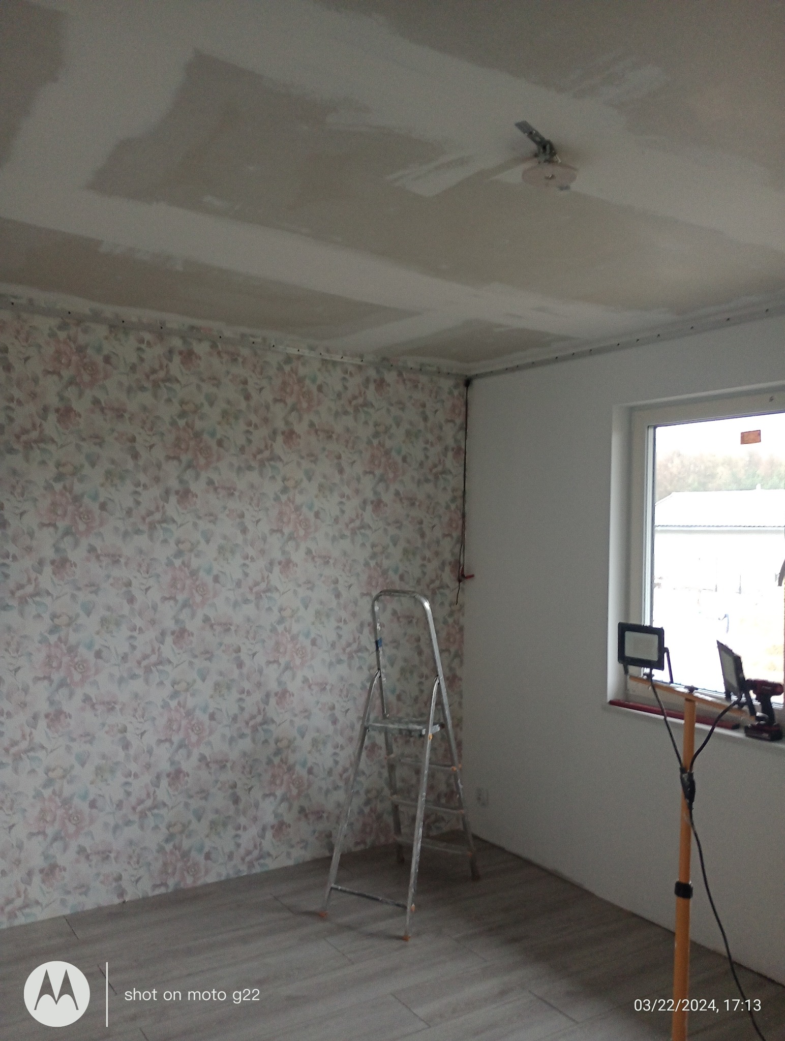 Pokój w trakcie remontu: ściana z tapetą w kwiaty, surowy sufit z widocznymi łączeniami płyt gipsowych, drabina i lampa oświetlająca okno. Widoczne przygotowania do malowania.