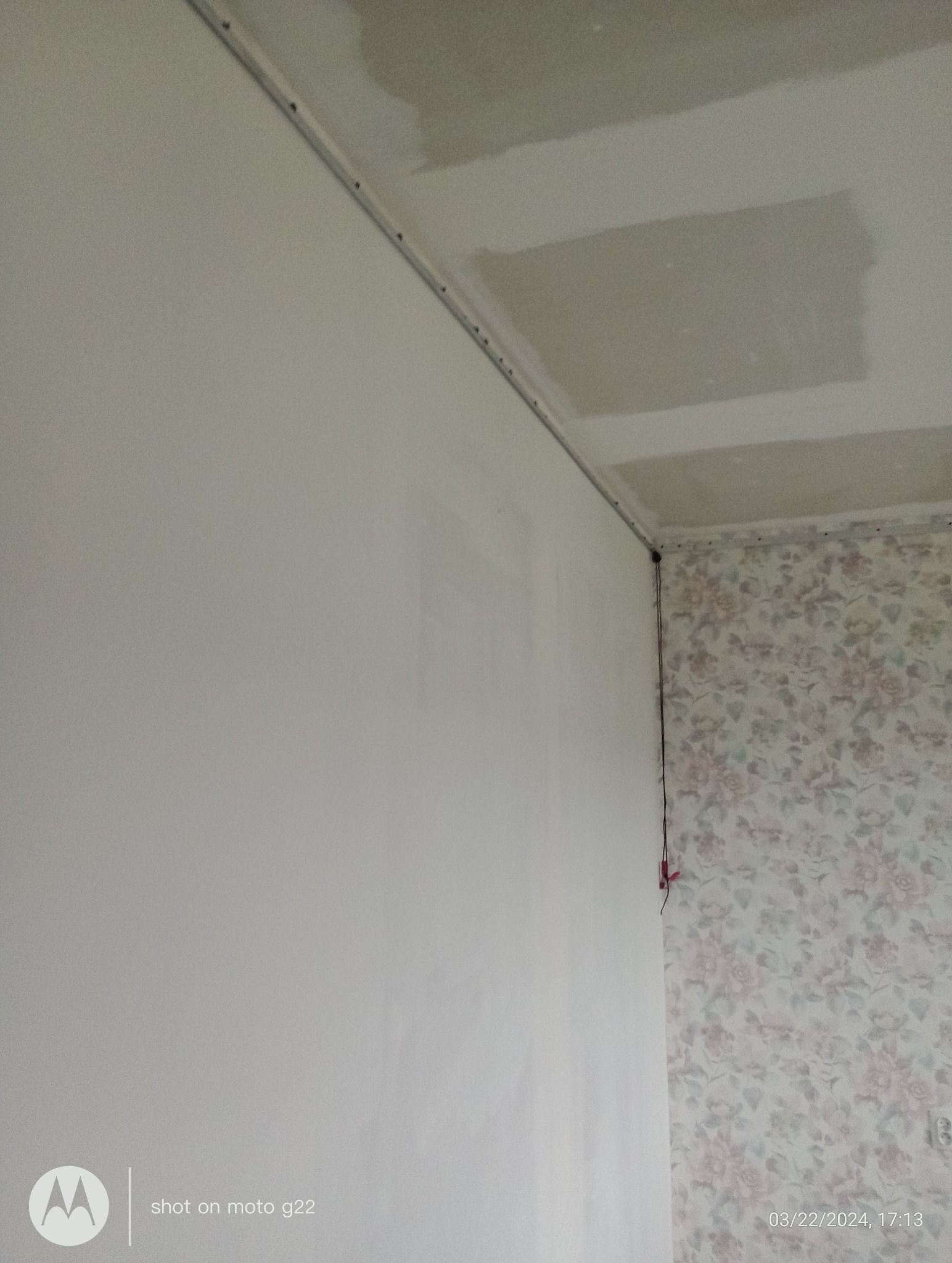 Przygotowana do malowania ściana z listwą sufitową i zagruntowanym sufitem. Fragment ściany z tapetą w kwiaty. Widoczne przewody elektryczne.