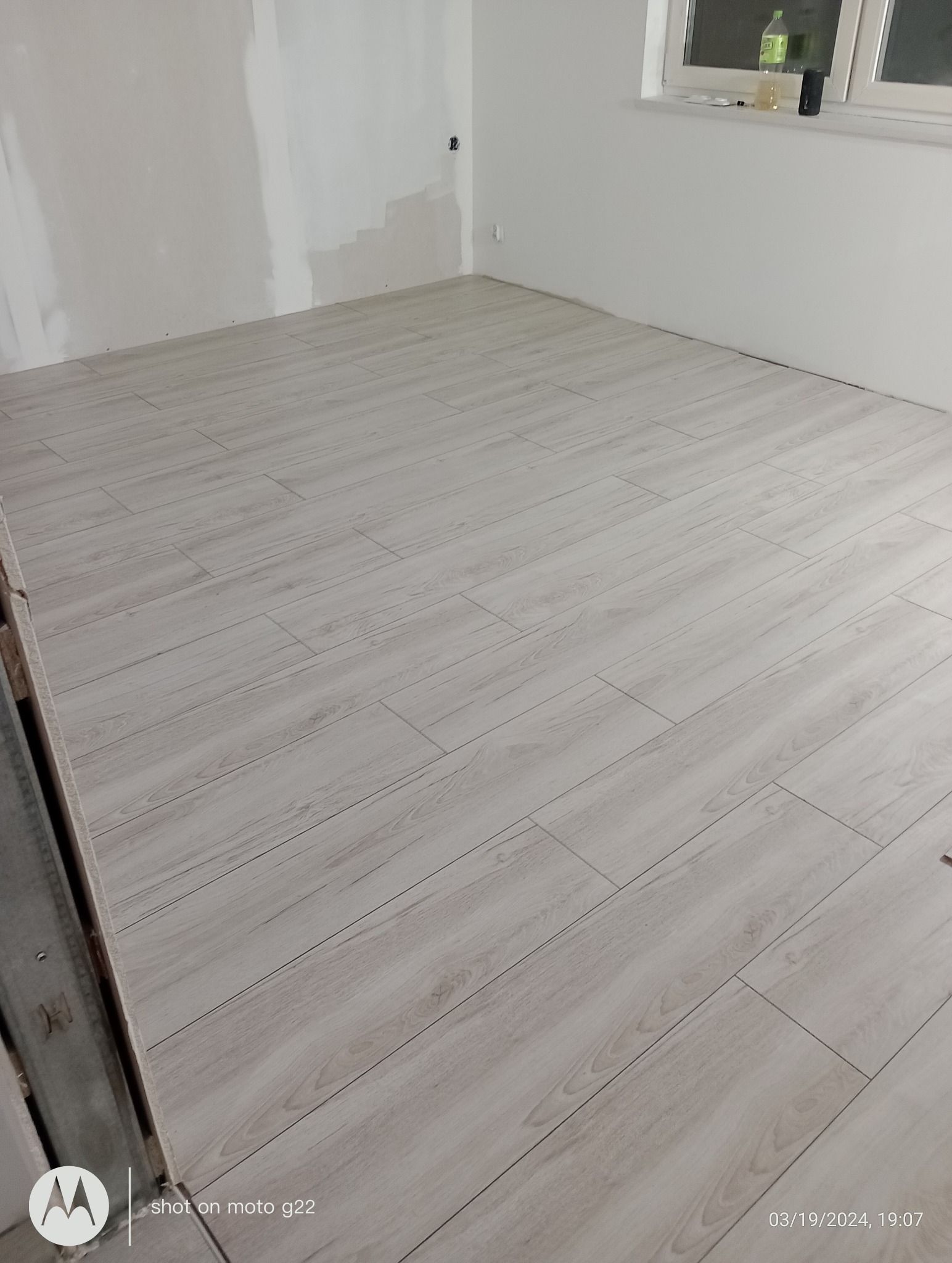 Ukończona podłoga z paneli imitujących drewno w jasnym odcieniu, w nowym pomieszczeniu z przygotowanymi ścianami do malowania. Widok na ułożone panele podłogowe.