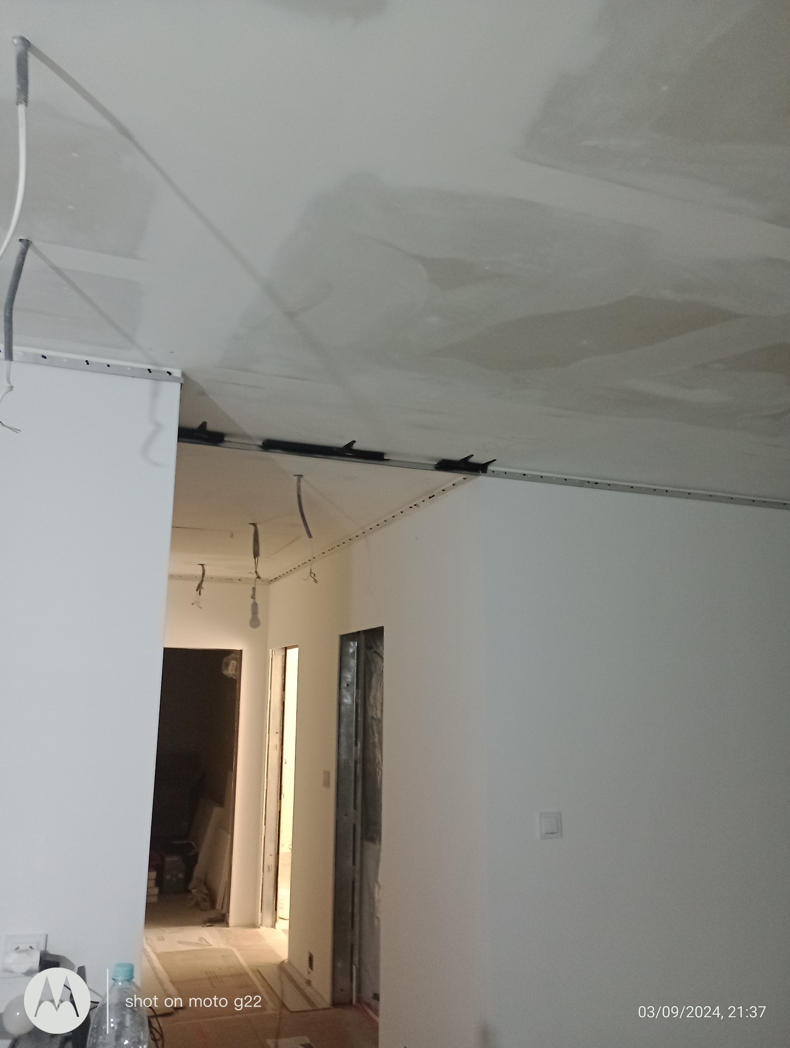 Wnętrze w trakcie remontu: białe ściany, sufit z płyt gipsowo-kartonowych, widoczne profile montażowe i instalacje elektryczne. Perspektywa z dołu, widok na korytarz.