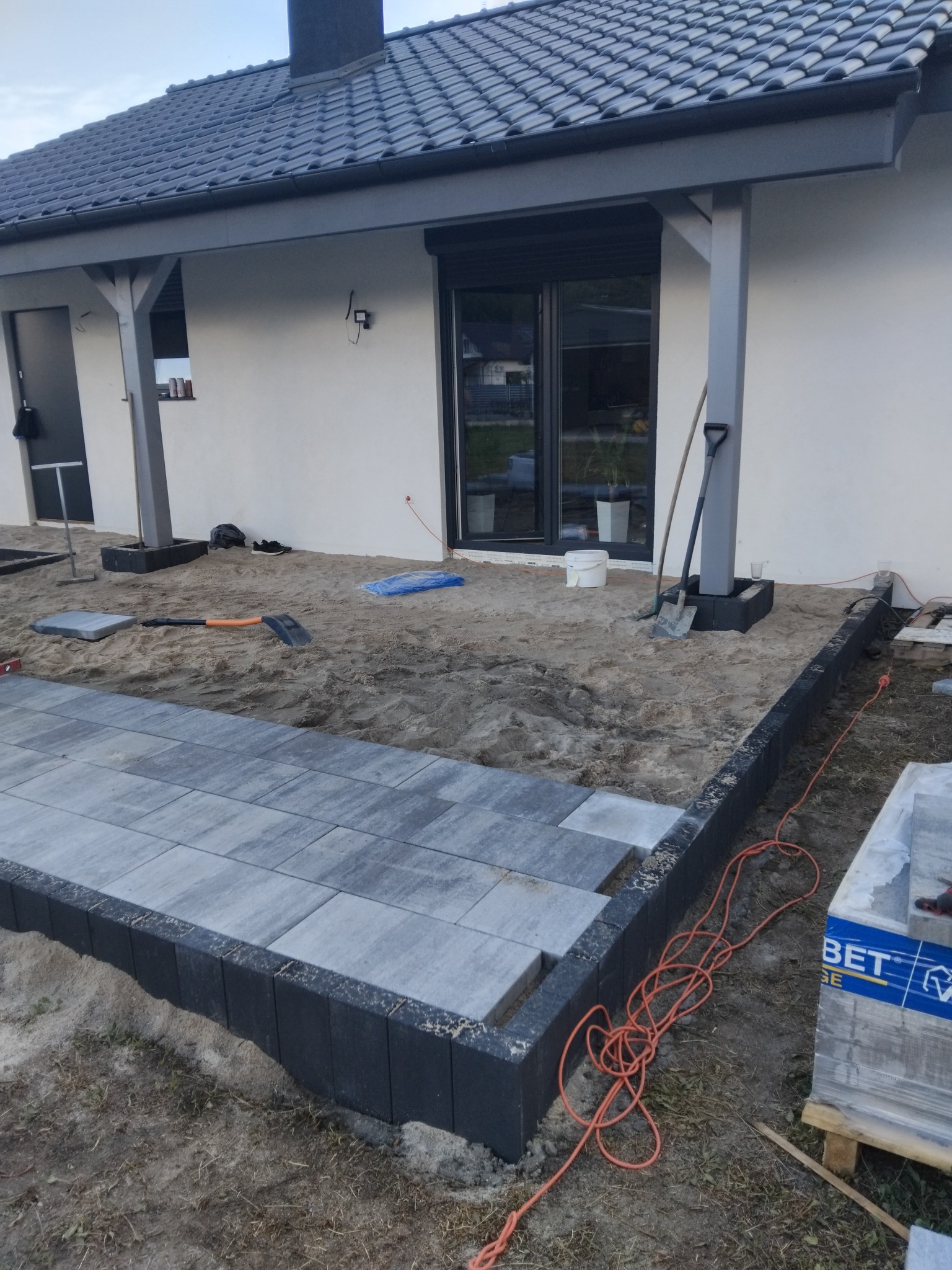 Układanie szarej kostki brukowej na podsypce piaskowej przy nowoczesnym domu z szarą stolarką okienną i ciemną dachówką. Widoczne narzędzia i materiały budowlane.