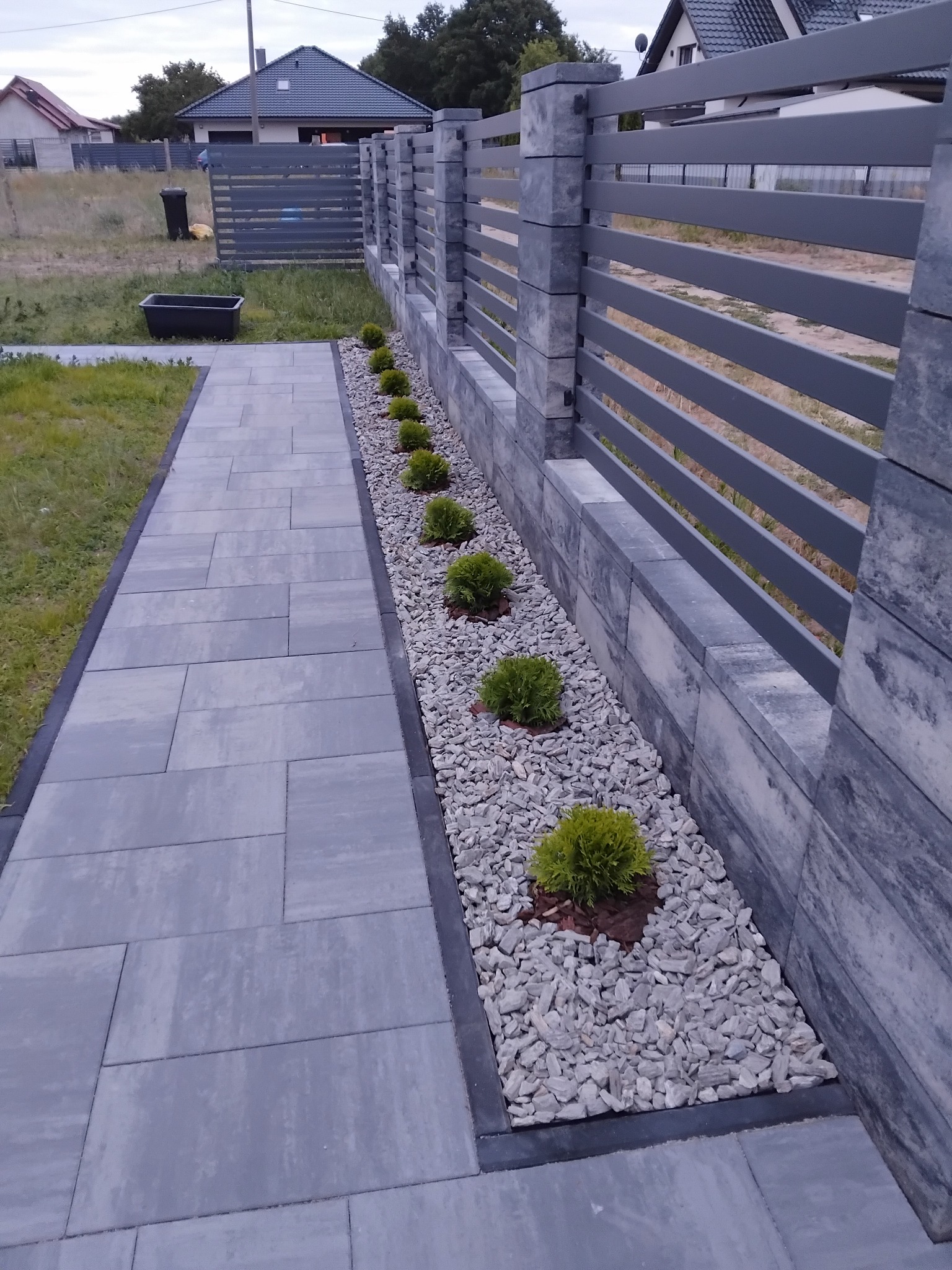 Szary chodnik z kostki brukowej, obok rabata z białym grysem i niskimi krzewami, oddzielona od posesji nowoczesnym, szarym ogrodzeniem panelowym.