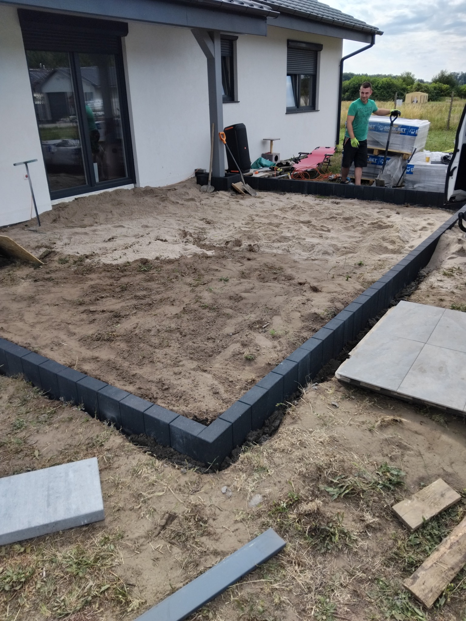 Przygotowanie terenu pod kostkę brukową: obrzeża ułożone, piasek wyrównany, paleta kostki w tle. Mężczyzna opiera się o łopatę, gotowy do dalszej pracy.