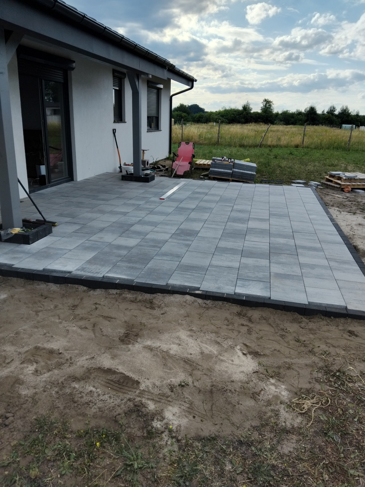 Ułożona, szara kostka brukowa na posesji domu jednorodzinnego z widocznymi narzędziami i paletami z kostką w tle. Widok na ogród i pochmurne niebo.