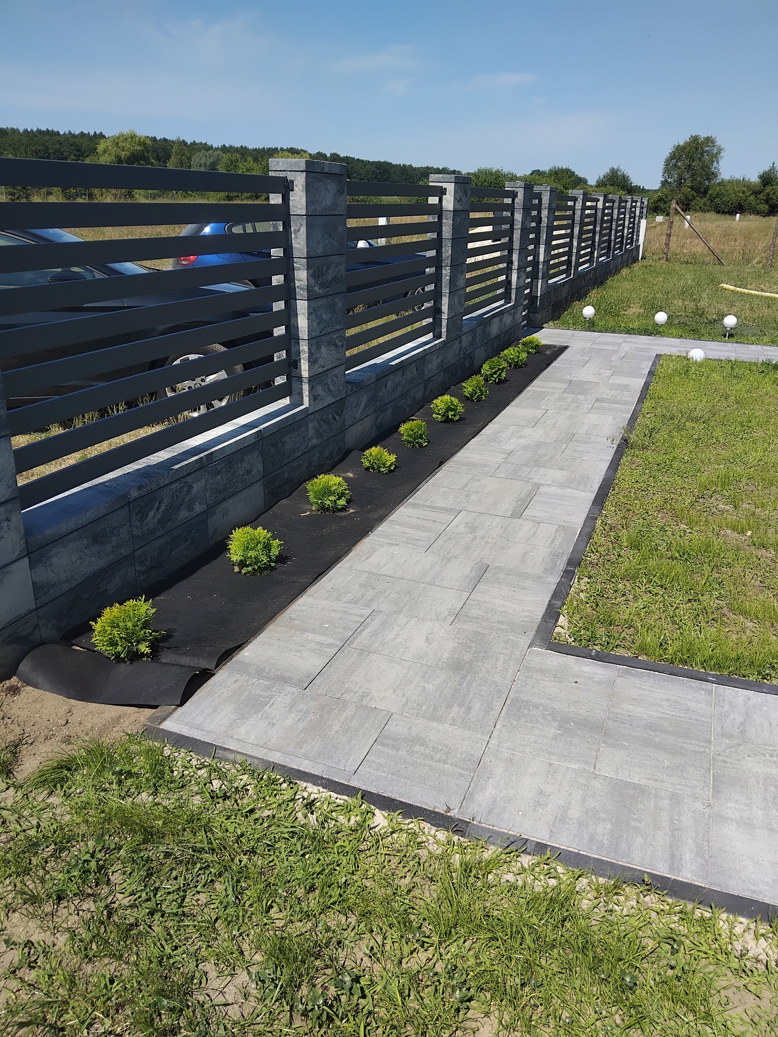 Nowoczesny chodnik z szarej kostki brukowej, obok rabata z niskimi krzewami i czarną agrowłókniną, w tle grafitowe ogrodzenie panelowe.