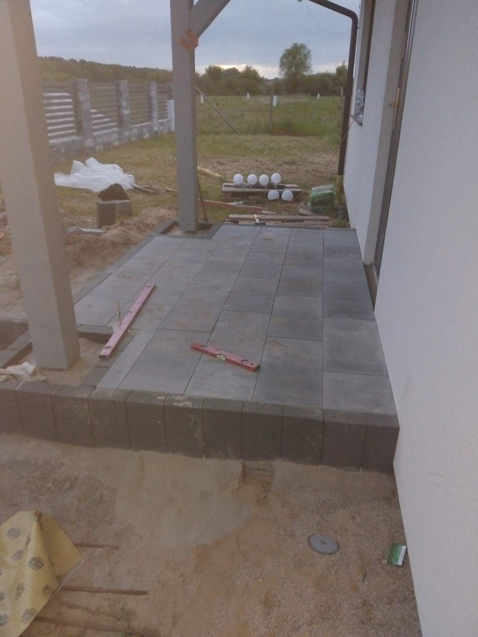 Układanie szarej kostki brukowej na tarasie przy domu, widoczne poziomice i elementy konstrukcyjne z drewna. Częściowo ułożona nawierzchnia.