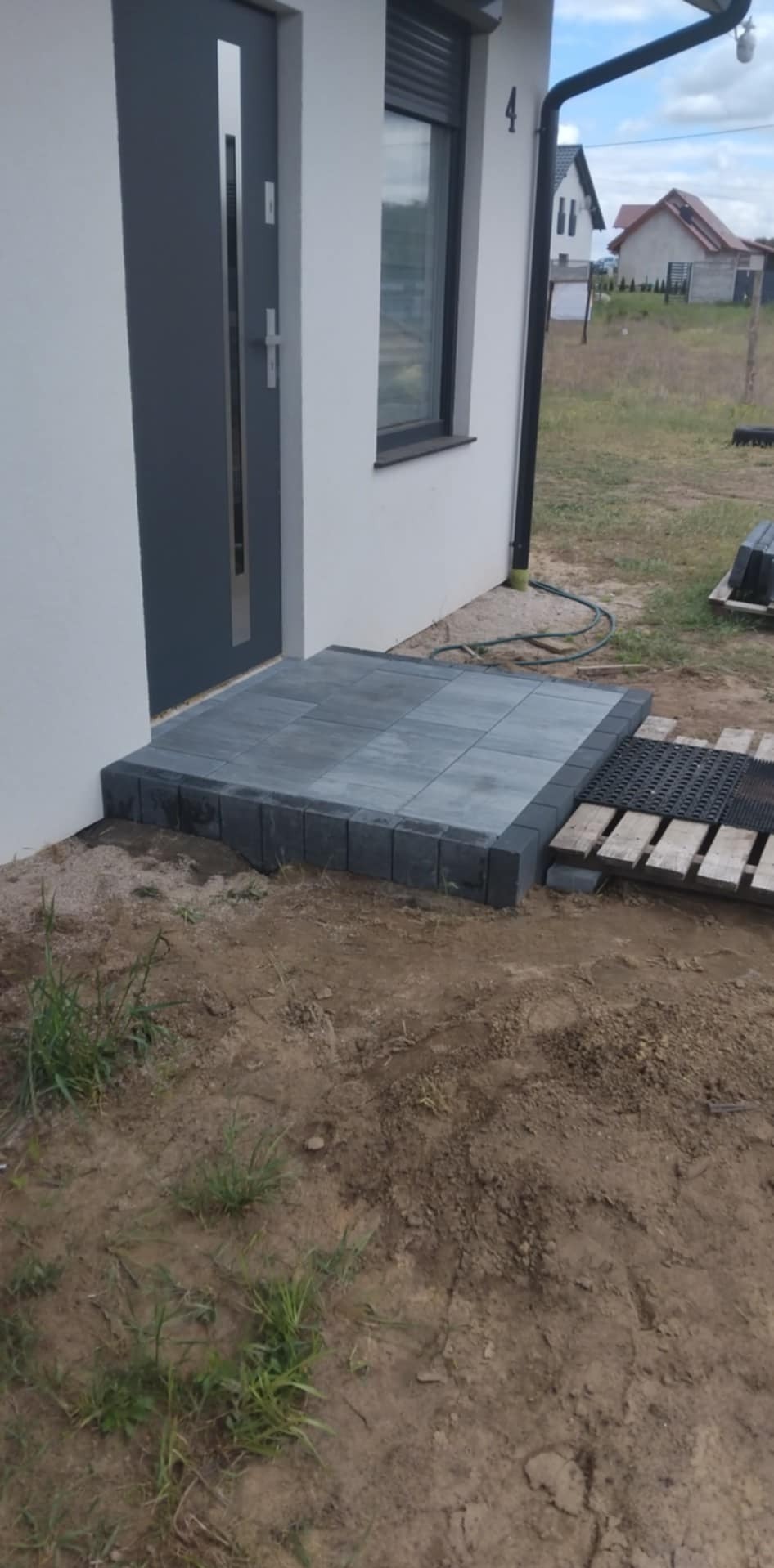 Ułożony, szary podest z kostki brukowej przed wejściem do domu z szarymi drzwiami i oknem z roletą. Widoczne palety i kostka brukowa w tle.