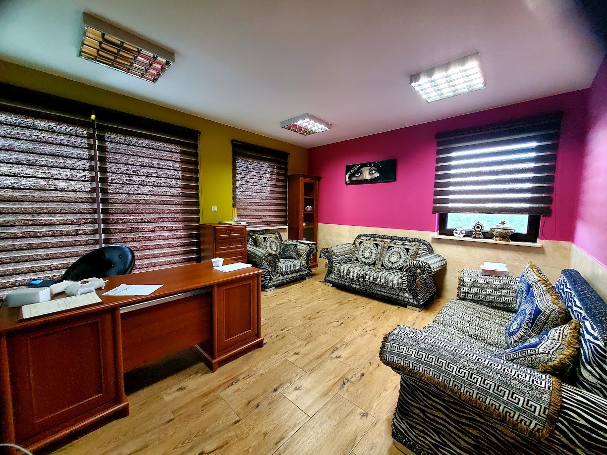 Gabinet z biurkiem, sofami w stylu Versace i obrazem oczu na różowej ścianie. Żaluzje poziome w oknach. Podłoga drewniana. Lampy sufitowe rastrowe.
