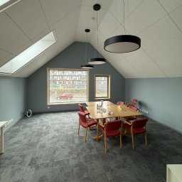 F.H.U AGNIESZKA STANIKOWSKA - Jasne, przestronne poddasze biurowe z szarymi ścianami, wykładziną i stołem konferencyjnym. Okna dachowe w skośnym suficie wpuszczają naturalne światło.