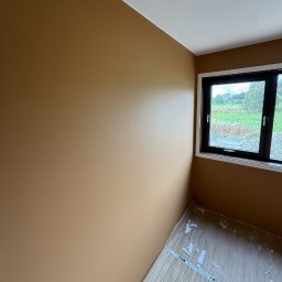 F.H.U AGNIESZKA STANIKOWSKA - Ściana w odcieniach brązu po malowaniu, widok na okno z widokiem na zieleń. Podłoga zabezpieczona kartonem. Świeżo pomalowane wnętrze.