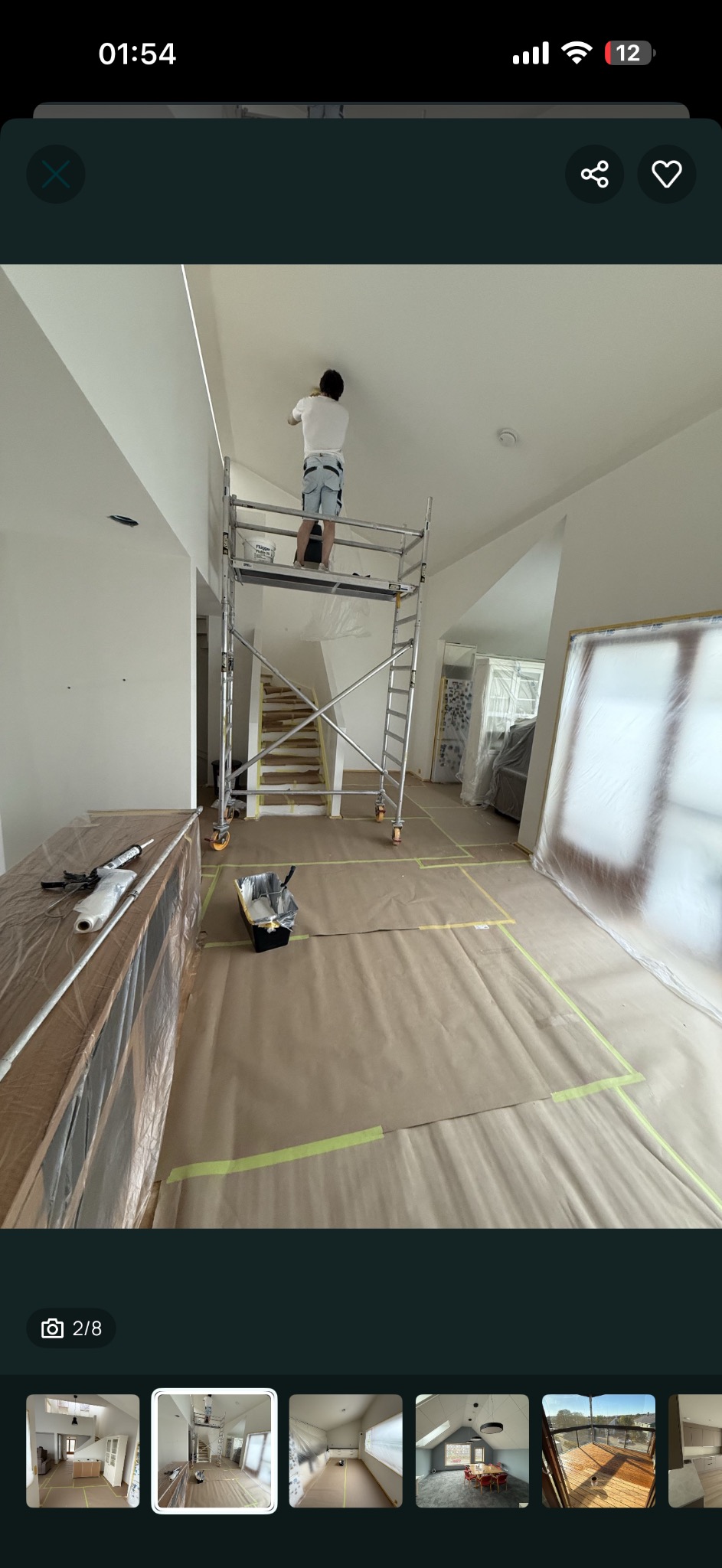 Pracownik na rusztowaniu maluje sufit w jasnym wnętrzu. Podłoga zabezpieczona papierem i taśmą. Widoczne schody i okna zabezpieczone folią. Etap przygotowania do malowania.