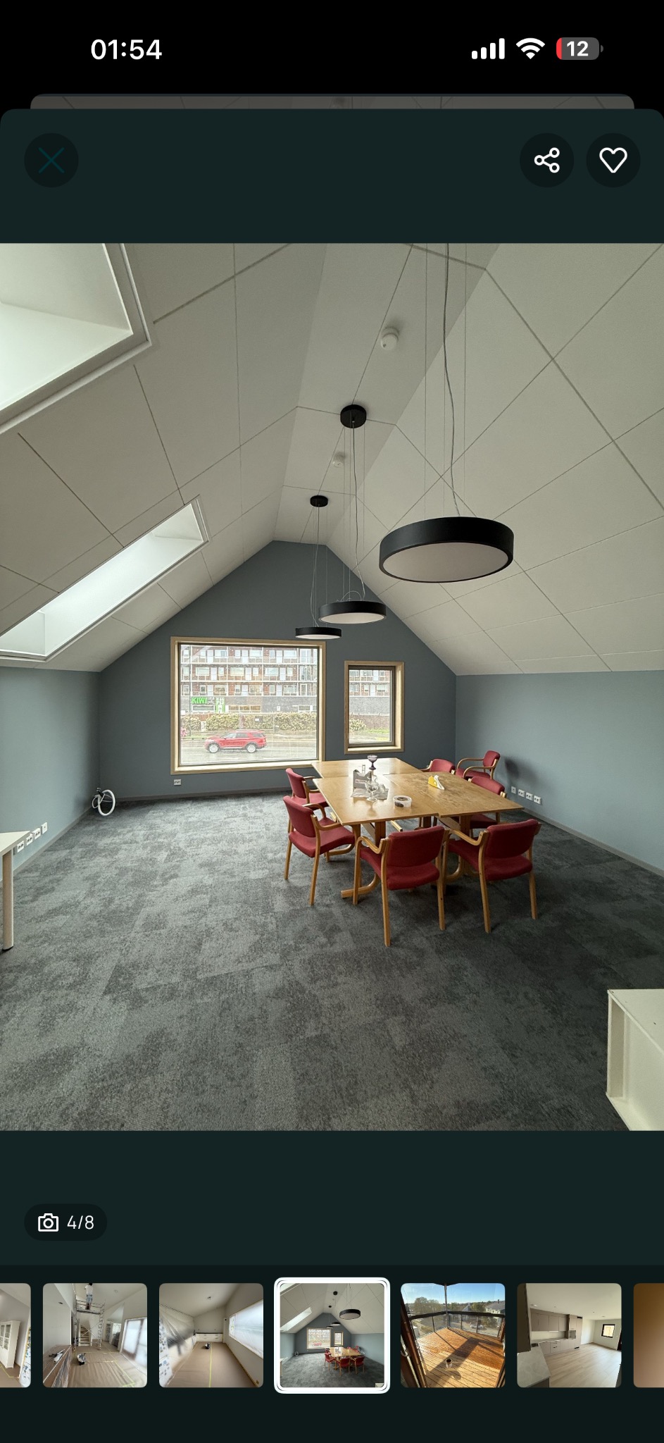 Jasne, przestronne poddasze biurowe z szarymi ścianami, wykładziną i stołem konferencyjnym. Okna dachowe w skośnym suficie wpuszczają naturalne światło.