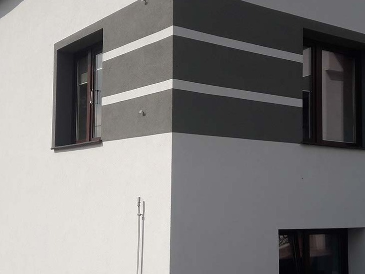 Fragment elewacji budynku z szarymi pasami i brązowymi oknami. Biała ściana kontrastuje z szarym narożnikiem. Widoczne detale wykończenia.