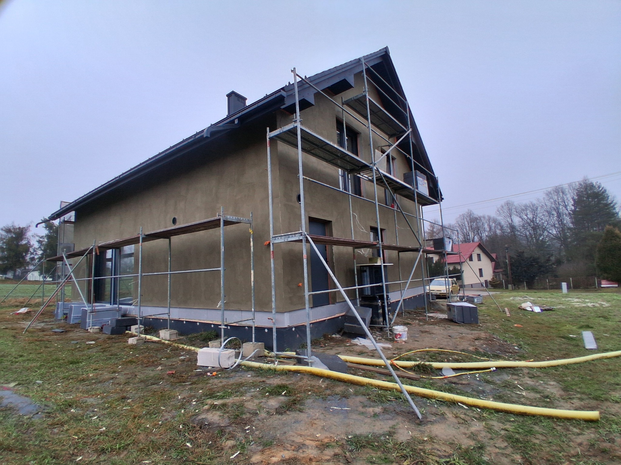 Dom jednorodzinny w trakcie tynkowania elewacji, widoczne rusztowania i przygotowane materiały. Szara, otynkowana ściana, czarne okna i dach. Pochmurne niebo.