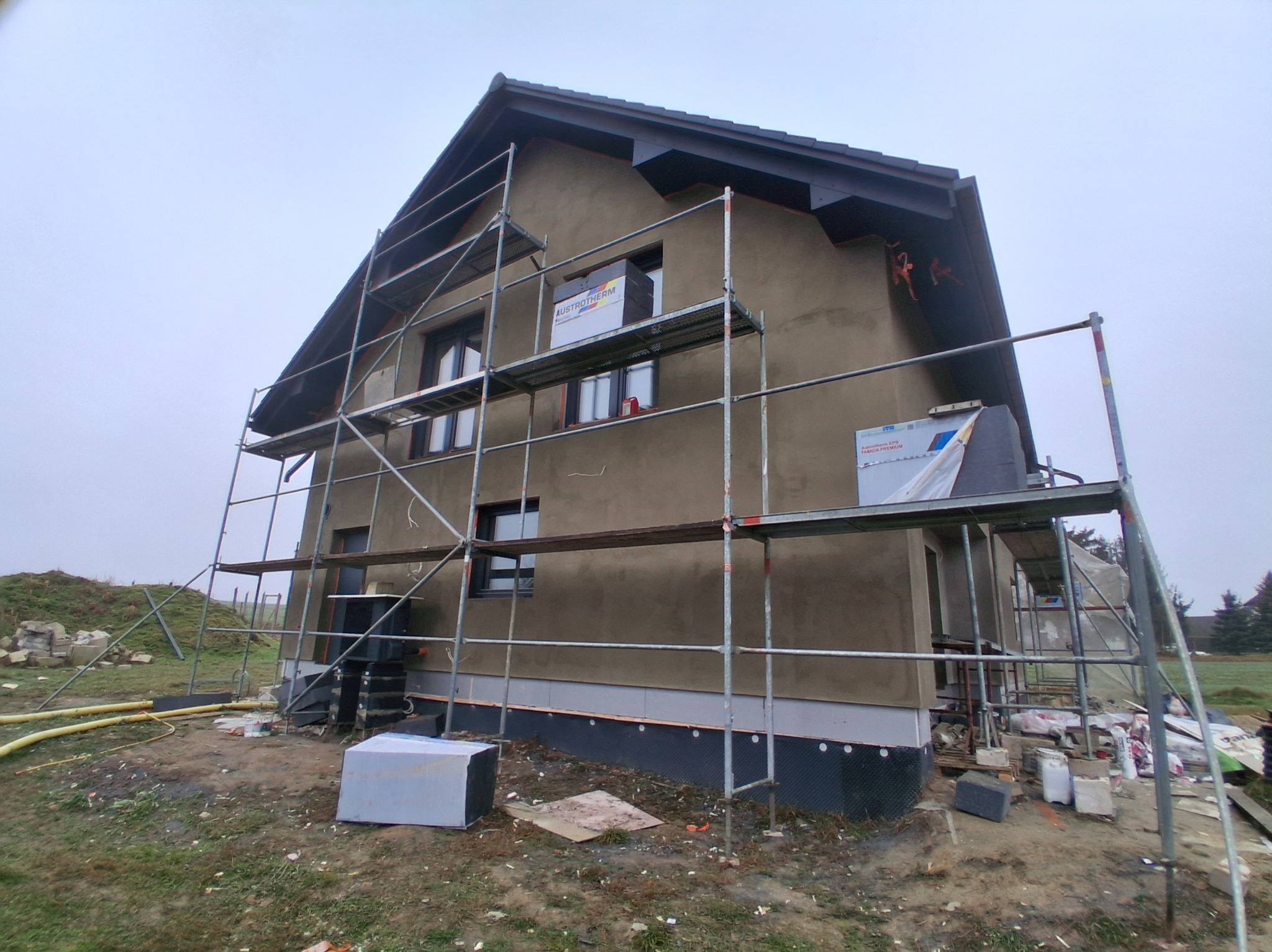 Dom w trakcie prac elewacyjnych: rusztowanie, ocieplenie częściowe, widoczne materiały izolacyjne. Świeżo otynkowana elewacja w kolorze beżowym.