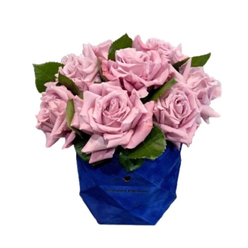 Bukiet pastelowych róż w geometrycznym, niebieskim wazonie. Kwiaty o delikatnym odcieniu fioletu, z zielonymi liśćmi, na białym tle. Dekoracja florystyczna.